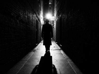 A mysterious man is lurking in a dark alley. A light is shining from behind him, so his face cannot be seen.