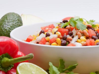 A white bowl of corn and bean salad surrounded by a red pepper, avocado, lime, and fresh cilantro.