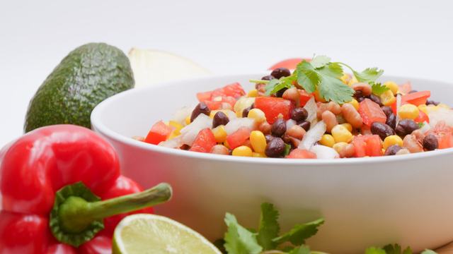 A white bowl of corn and bean salad surrounded by a red pepper, avocado, lime, and fresh cilantro.