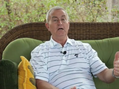 A man wearing a striped polo shirt sits on a wicker couch with green cushions and yellow pillows. He's gesturing with his hand while speaking.
