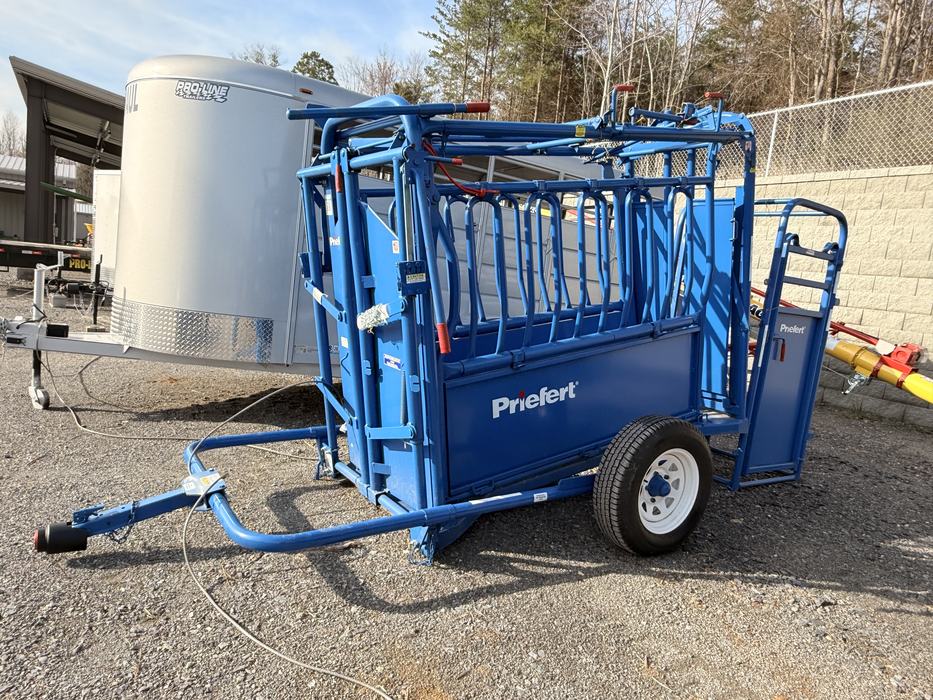 A bright blue Priefert squeeze chute with an attached palpation cage is mounted on a mobile trailer frame with white wheels.