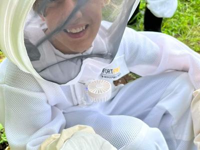 A smiling person in a white beekeeping suit and veil kneels outdoors, gently holding a small bee on their gloved hand.