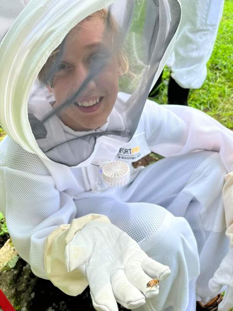 A smiling person in a white beekeeping suit and veil kneels outdoors, gently holding a small bee on their gloved hand.