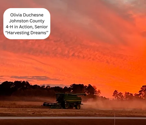 A combine harvester moves across a field at sunset, kicking up dust beneath a dramatic orange-red sky, with dark tree silhouettes along the horizon.