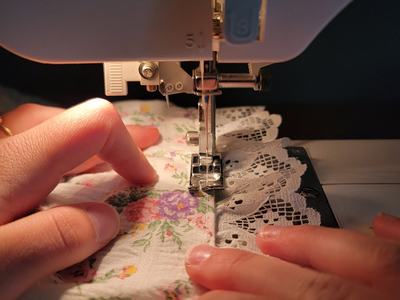 Close-up of hands guiding floral fabric with lace trim under the needle of a sewing machine, stitching along the fabric's edge.