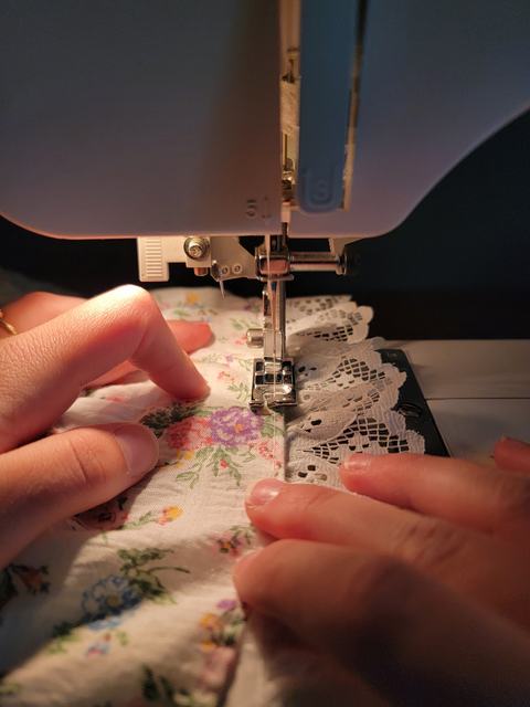 Close-up of hands guiding floral fabric with lace trim under the needle of a sewing machine, stitching along the fabric's edge.