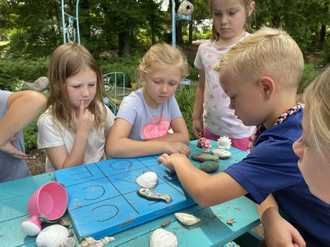 Children gathered around a turquoise picnic table outdoors, playing tic-tac-toe on a painted wooden board using shells and painted rocks as markers; a boy places a piece while others watch.