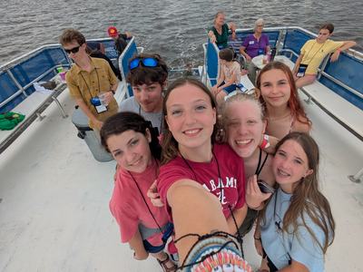 A group of smiling teens take a selfie on a boat’s deck, the photographer’s arm and bracelets in the foreground, wake trailing behind them across calm water under a cloudy sky.