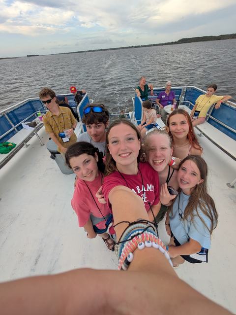 A group of smiling teens take a selfie on a boat’s deck, the photographer’s arm and bracelets in the foreground, wake trailing behind them across calm water under a cloudy sky.