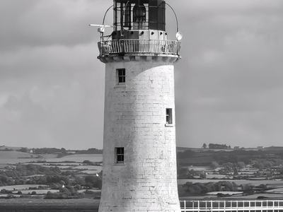 Black-and-white photo of a solitary cylindrical lighthouse rising from the sea, topped by a lantern and gallery, linked to shore by a narrow pier beneath a cloudy sky.