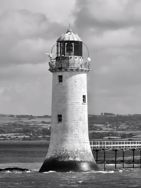 Black-and-white photo of a solitary cylindrical lighthouse rising from the sea, topped by a lantern and gallery, linked to shore by a narrow pier beneath a cloudy sky.
