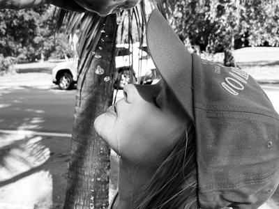 Black-and-white photo of a person in a baseball cap tilting their head back to drink coconut water held by another hand, with a palm tree and parked car in the background.