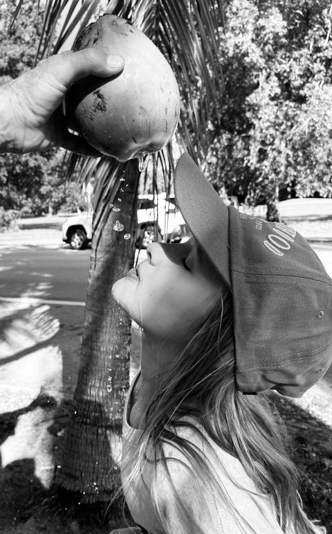 Black-and-white photo of a person in a baseball cap tilting their head back to drink coconut water held by another hand, with a palm tree and parked car in the background.