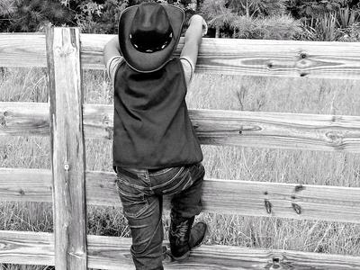 Black-and-white photo of a child wearing a cowboy hat, vest and boots climbing a wooden fence, leaning to peer over at a grassy field and trees beyond.
