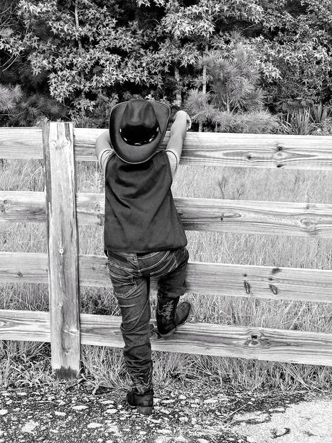 Black-and-white photo of a child wearing a cowboy hat, vest and boots climbing a wooden fence, leaning to peer over at a grassy field and trees beyond.