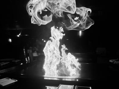 Black-and-white photo of tall, dancing flames rising from a teppanyaki-style grill in a restaurant, with a plate and dipping dishes in the foreground.