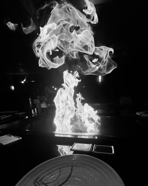 Black-and-white photo of tall, dancing flames rising from a teppanyaki-style grill in a restaurant, with a plate and dipping dishes in the foreground.