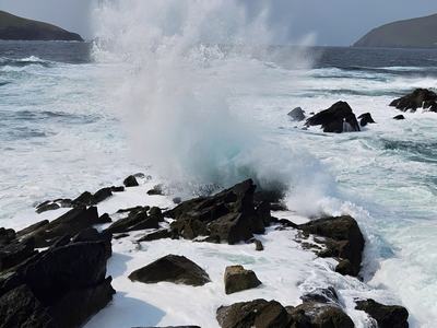 Ocean waves slam into jagged black rocks along a rugged coast, sending a towering spray of white foam under a cloudy sky.