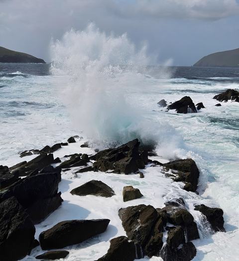 Ocean waves slam into jagged black rocks along a rugged coast, sending a towering spray of white foam under a cloudy sky.