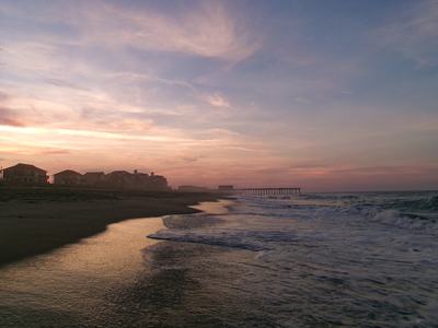 Pastel sunset over a quiet beach: gentle waves lap a reflective shoreline, silhouetted beach houses along the coast and a long pier stretching into the calm sea.