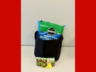 Black 5-gallon grow bag filled with potting mix and a bag of potting soil, displayed with vegetable seed packets and a small planting tool provided to participants in the Seed to Supper gardening program