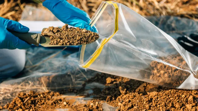 A person wearing blue gloves uses a hand trowel to collect soil and place it into a clear plastic sample bag in an outdoor setting.