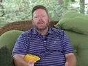 A man with a beard sits on a wicker sofa with green cushions outdoors, wearing a blue and pink striped polo shirt and gesturing with his hands.