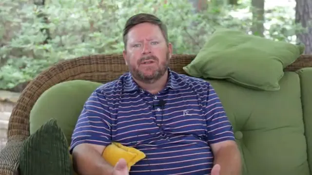 A man with a beard sits on a wicker sofa with green cushions outdoors, wearing a blue and pink striped polo shirt and gesturing with his hands.