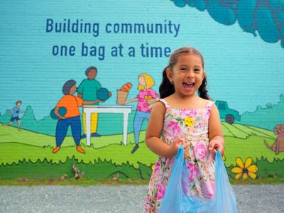 Female child carrying bag of food in front of a painting on wall that says "building community one bag at a time".