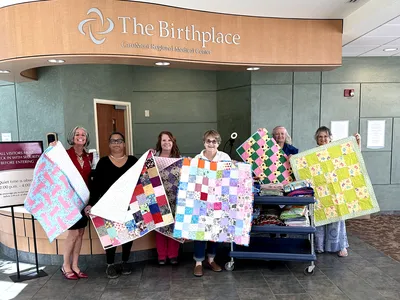 Six women, three ECA members, and three caregivers stand under the awning of The Birthplace at CaroMont Regional Medical Center. They are each holding up a donated baby quilt made by members of the ECA Quiltmakers club.