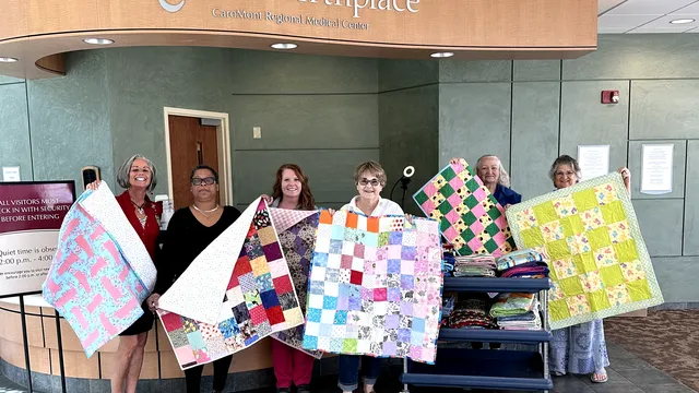 Six women, three ECA members, and three caregivers stand under the awning of The Birthplace at CaroMont Regional Medical Center. They are each holding up a donated baby quilt made by members of the ECA Quiltmakers club.