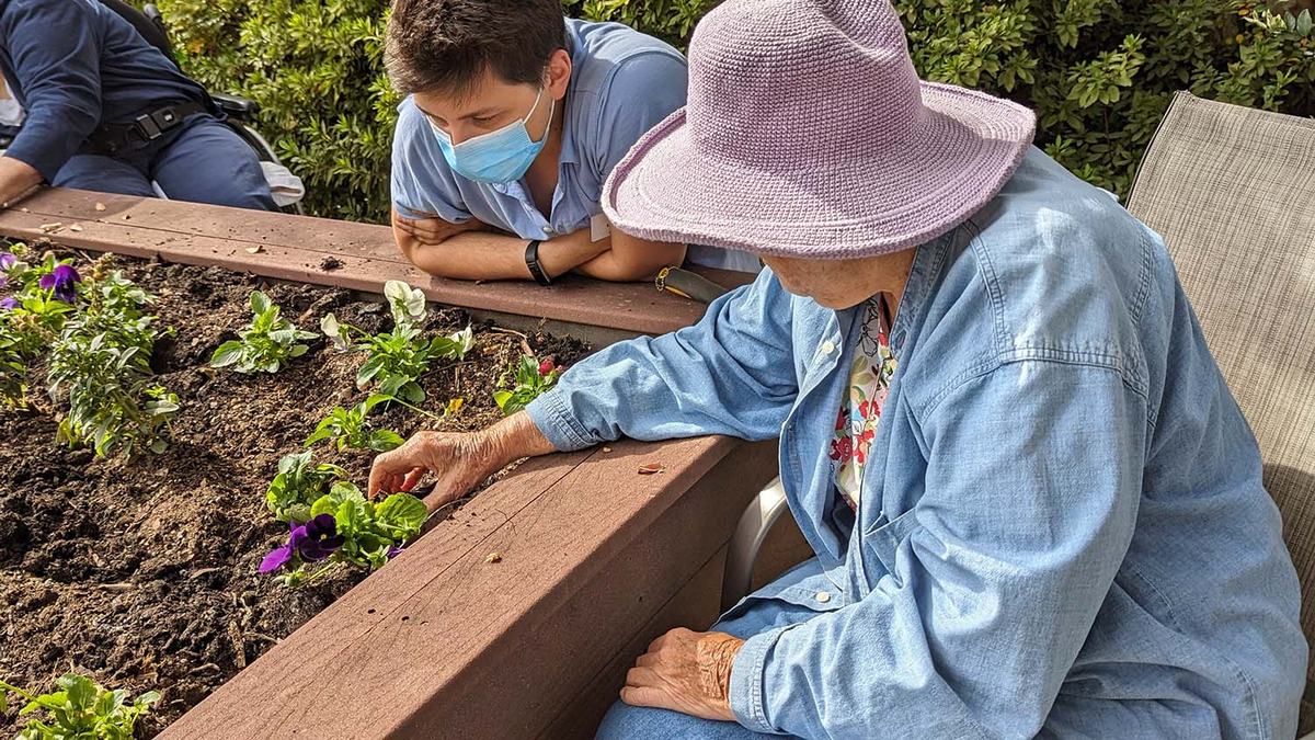 A seated elderly woman digs in a raised garden bed while a young man looks on