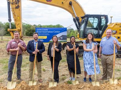 L t R: Aaron Moore, Andrew Baucom, Jessica King, Samara Gibson, Ashlin Austin, Keith Walters; all stand with a shovel at a construction site with a photo of the future of the FIC in the background