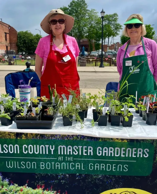 Two Wilson County Master Gardener volunteers posing in front of informational booth with Master Gardener and Wilson Botanical Garden resources.