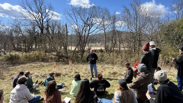 Specialist in red baseball cap talking to group of people in a field adjacent to a river