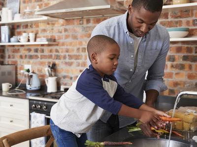A father helping his young son wash fresh produce, including carrots and corn, at their kitchen sink.