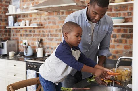 A father helping his young son wash fresh produce, including carrots and corn, at their kitchen sink.