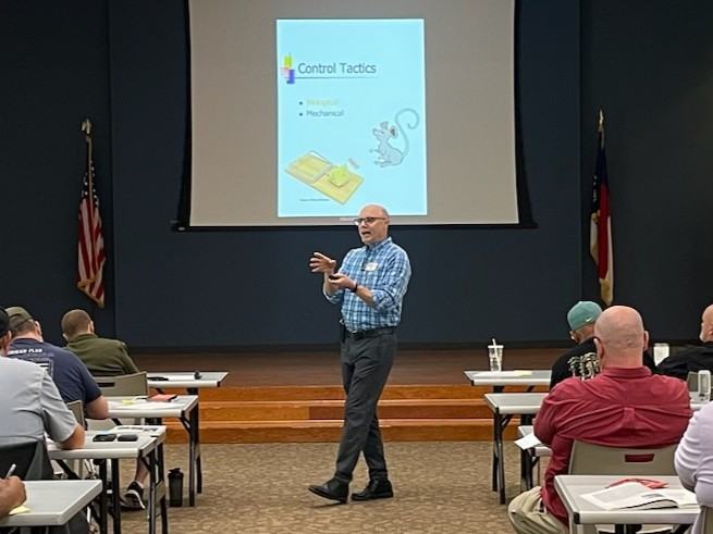 Wayne Buhler teaching the Pesticide Safety School participants while walking and gesturing at the front of the classroom.