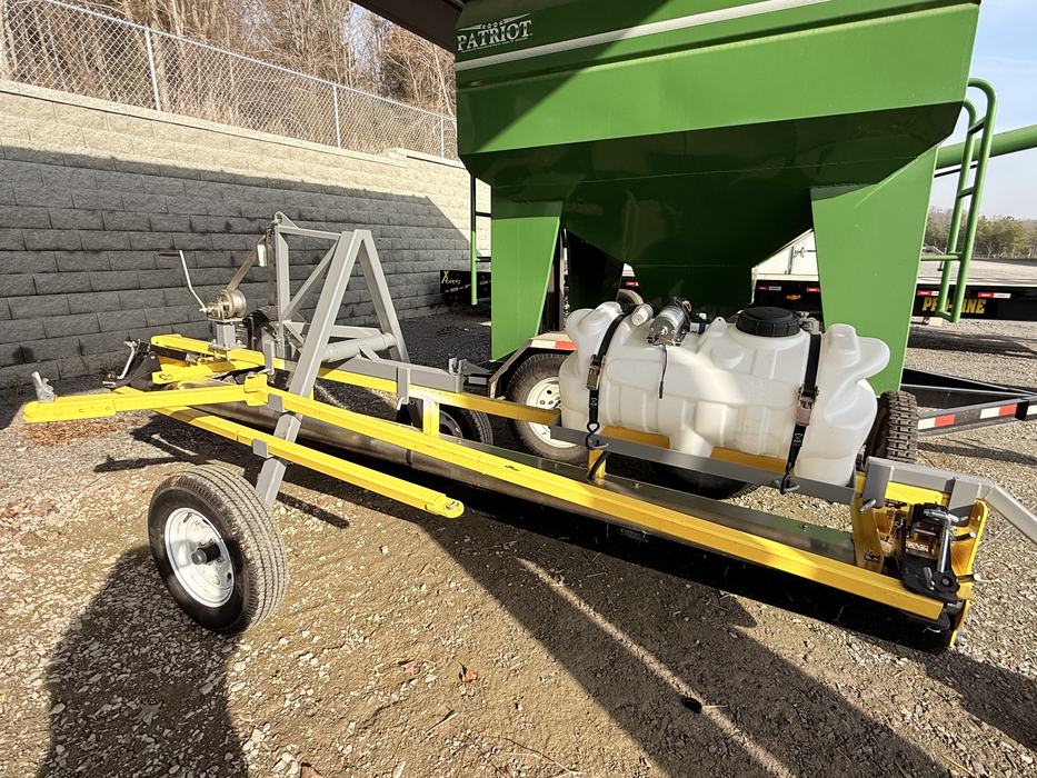 A yellow and gray weed wiper attachment on a wheeled trailer frame, featuring a white liquid tank secured with black straps. It is parked on gravel in front of a large green Patriot agricultural bin.
