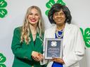 A young woman wearing a green blazer stands next to a woman wearing a white blazer who is holding an award plaque.