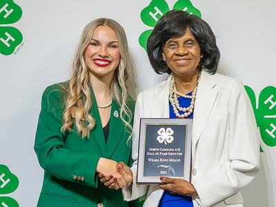 A young woman wearing a green blazer stands next to a woman wearing a white blazer who is holding an award plaque.