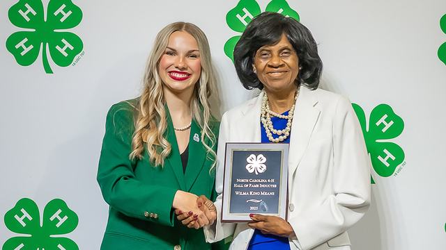 A young woman wearing a green blazer stands next to a woman wearing a white blazer who is holding an award plaque.