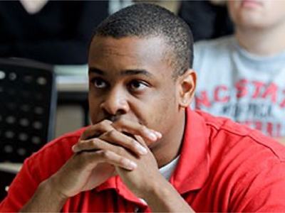 A young man in red shirt listens with hands folded under his chin during a class.