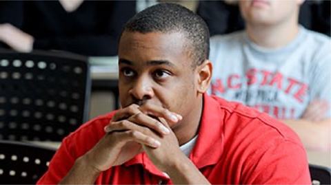 A young man in red shirt listens with hands folded under his chin during a class.
