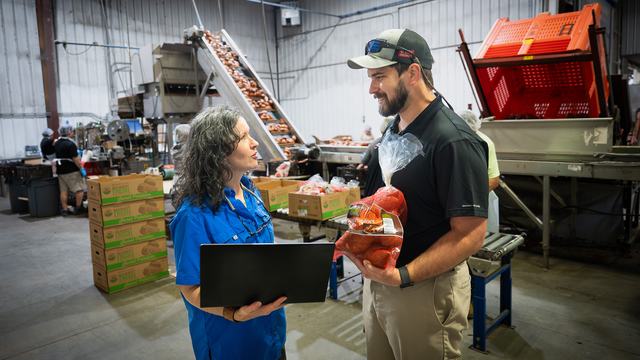 An Extension agent holds a bag of sweetpotatoes while talking with a farmer holding a laptop at their food processing facility.