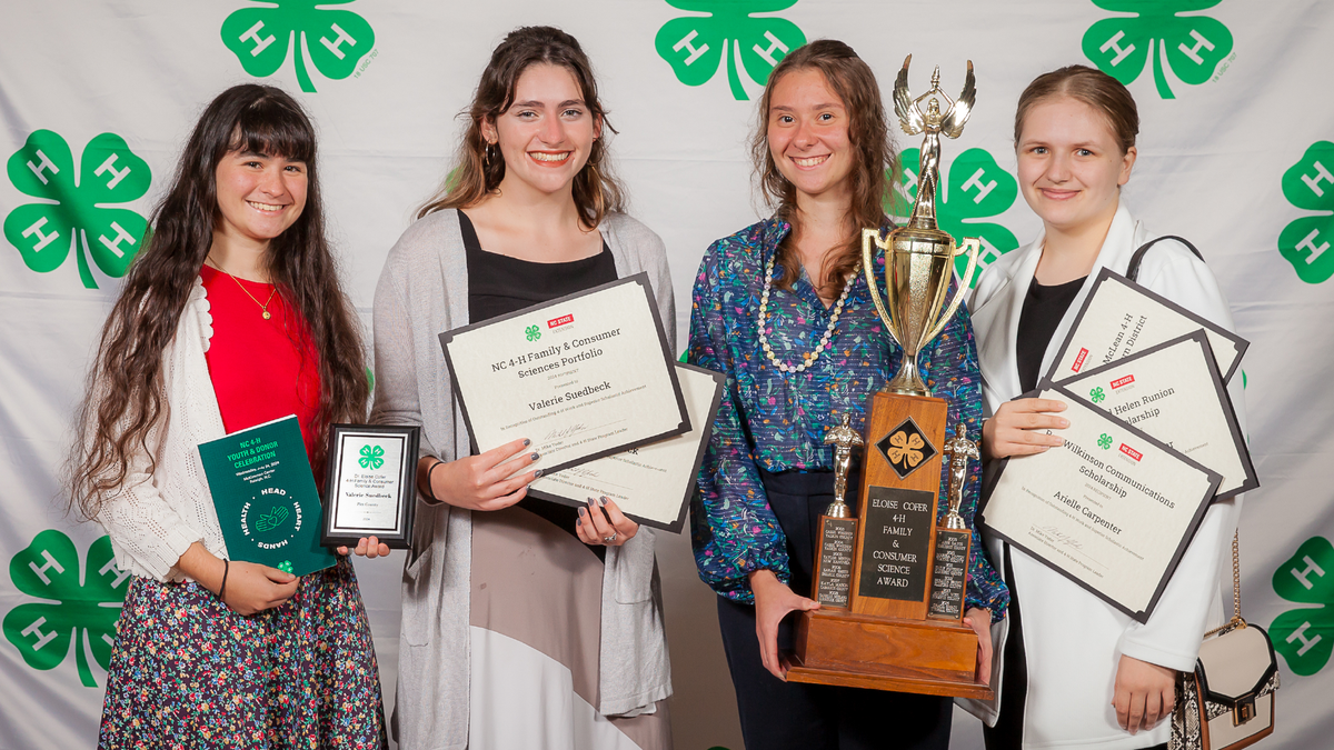 Four 4-H members in front of a 4‑H backdrop, holding certificates, plaques and a large trophy at the 2024 NC 4-H Youth and Donor event during 4-H Congress.