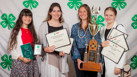 Four 4-H members in front of a 4‑H backdrop, holding certificates, plaques and a large trophy at the 2024 NC 4-H Youth and Donor event during 4-H Congress.