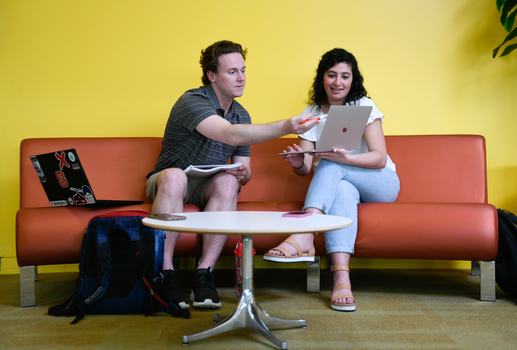 Two young people-one male, one female-sitting on a couch.  One holding a computer with the other pointing someting out on the computer.