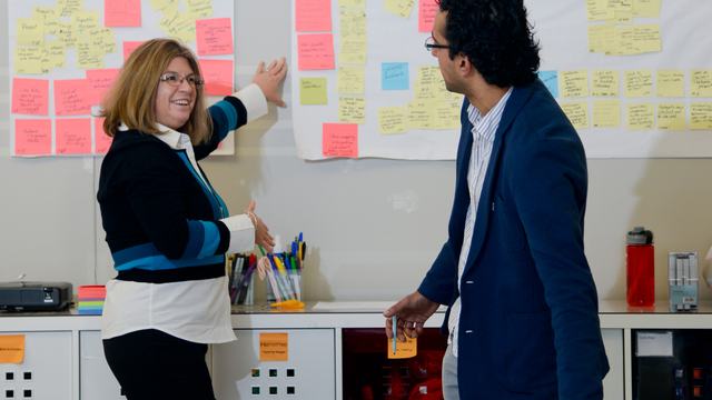 A woman points to sticky, colorful note among many across a white wall as a man over a table grabs a new sticky note to add.
