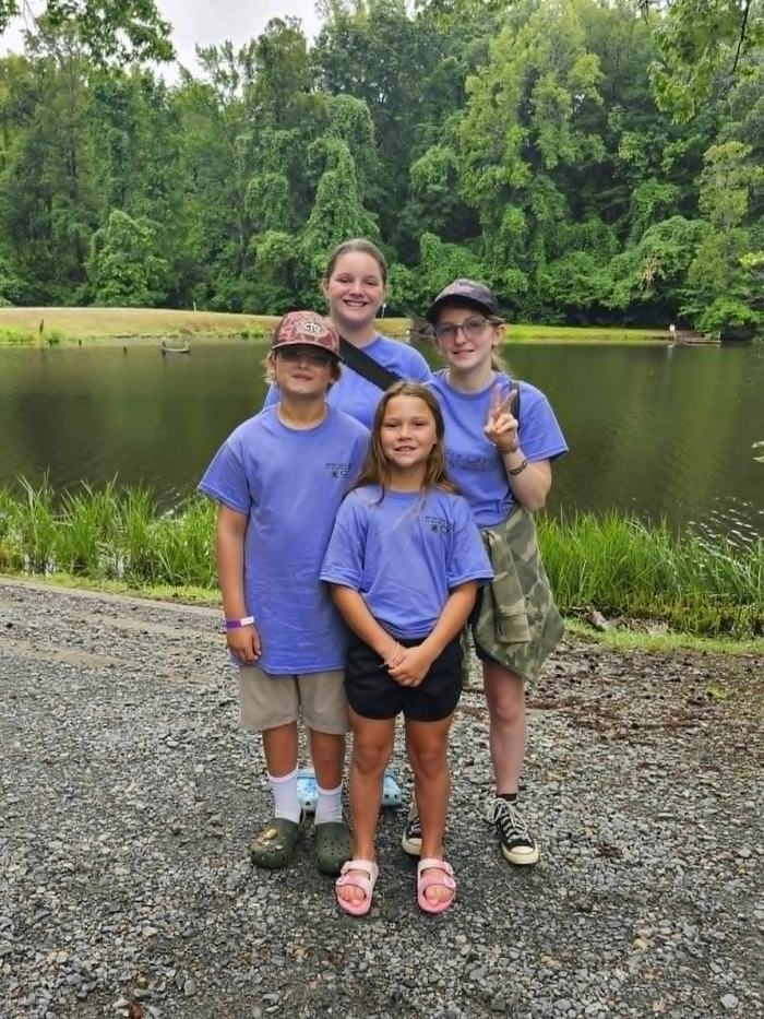 Four children wearing matching light blue t-shirts pose together on a gravel path in front of a calm pond surrounded by dense green trees. The group includes two older girls in the back and a younger boy and girl standing in front.